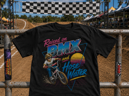 A black graphic t-shirt featuring a colorful retro design with the text 'Raised on BMX and Hose Water' and an illustration of a BMX rider, displayed on a metal fence at an outdoor dirt track event.